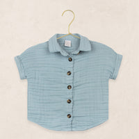 Lou Lou & Company Cloud Muslin Shirt in Steel, a soft blue button-front short-sleeve shirt on a hanger