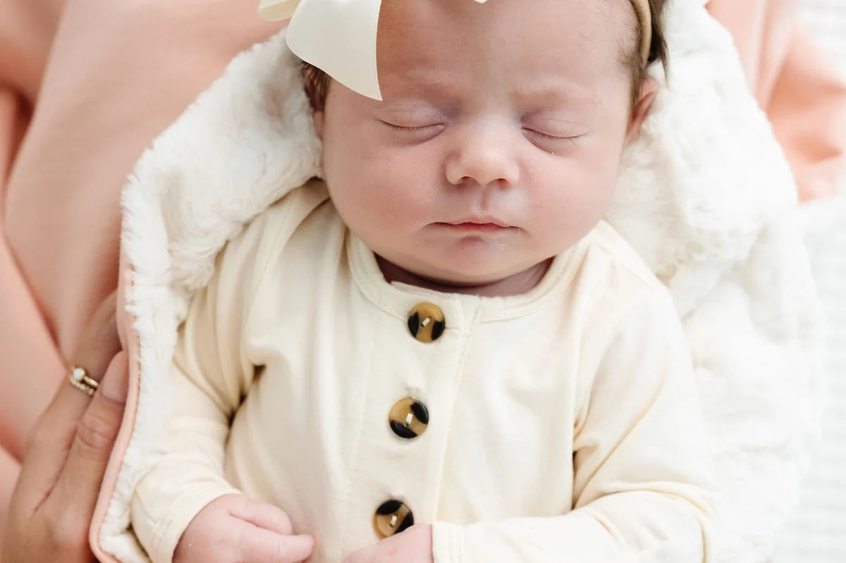 Newborn with a cream bow sleeping on the sherpa side of the Lou Lou & Company Cozy Blanket in Ballet Pink