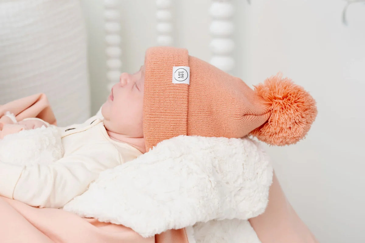 Newborn in a coral pom hat wrapped in the Lou Lou & Company Cozy Blanket in Ballet Pink