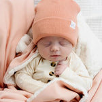 Sleeping newborn in a coral beanie snuggled in the Lou Lou & Company Cozy Blanket in Ballet Pink