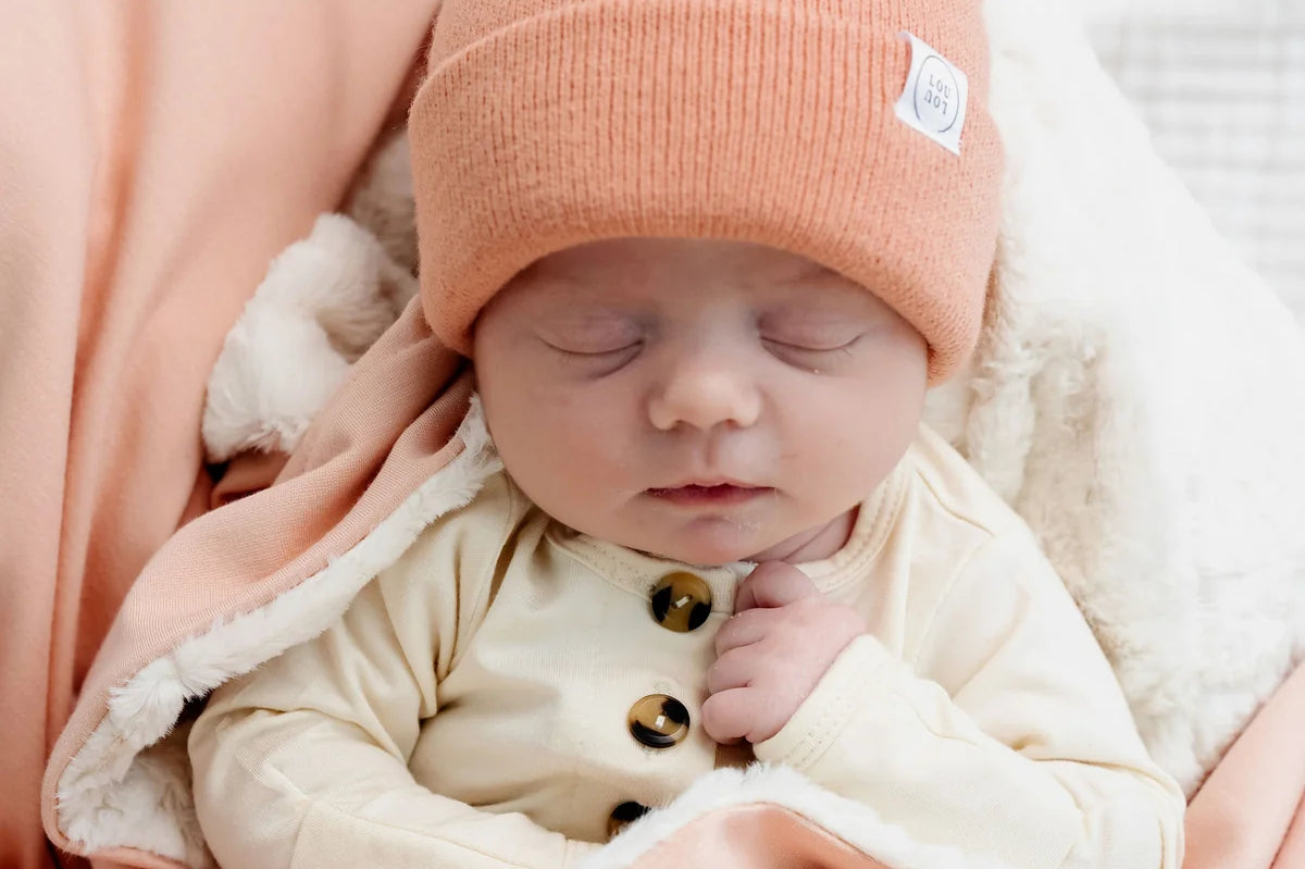 Sleeping newborn in a coral beanie snuggled in the Lou Lou & Company Cozy Blanket in Ballet Pink