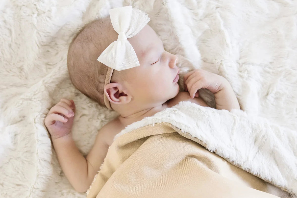Newborn with a white bow sleeping under the Lou Lou & Company Cozy Blanket in Latte on sherpa