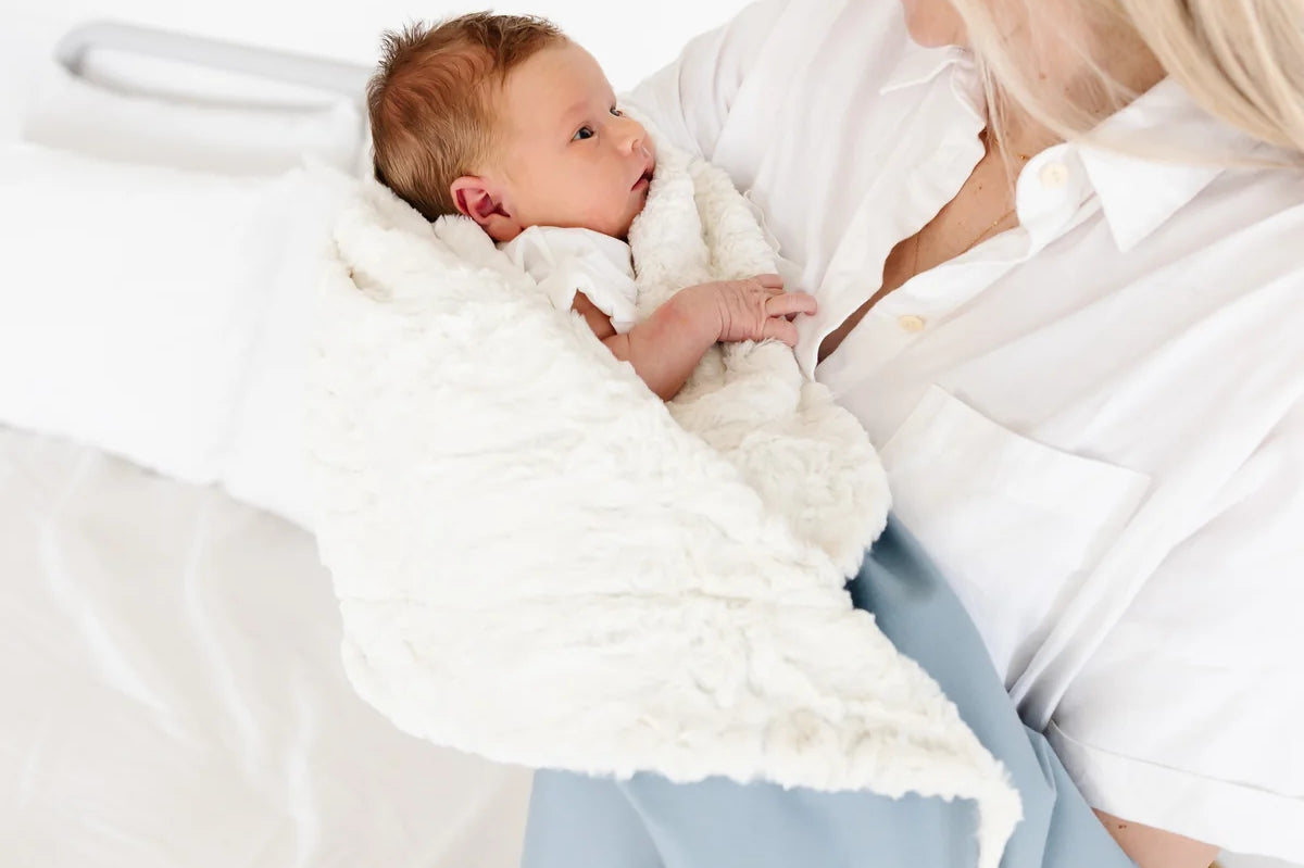 Mother holding a newborn wrapped in the Lou Lou & Company Cozy Blanket in Steel blue, sherpa side out