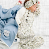Newborn in a floral romper and headband lying beside the Lou Lou & Company Cozy Blanket in Steel blue