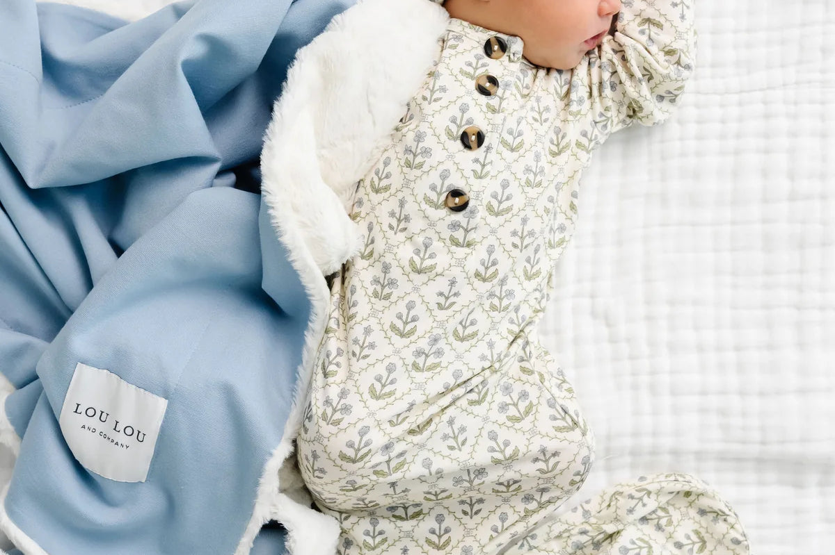 Newborn in a floral romper and headband lying beside the Lou Lou & Company Cozy Blanket in Steel blue