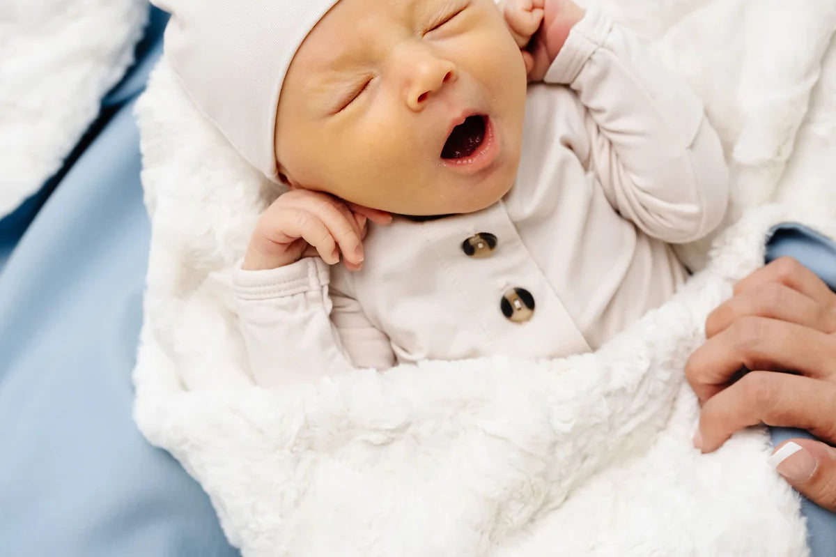 Yawning newborn in a white hat wrapped in the Lou Lou & Company Cozy Blanket in Steel blue