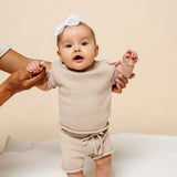 Baby with a white bow held up in the Lou Lou & Company Knit Tee and Shorts in Tan