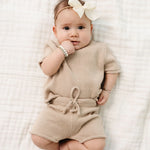 Baby with a white bow lying on a blanket in the Lou Lou & Company Knit Tee and Shorts in Tan