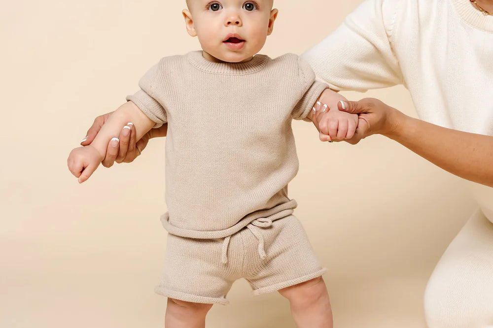 Baby standing with help wearing the Lou Lou & Company Knit Tee and Shorts in Tan