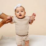 Baby with a white bow held up wearing the Lou Lou & Company Knit Shorts and Tee in Tan