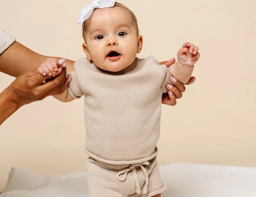 Baby with a white bow held up wearing the Lou Lou & Company Knit Shorts and Tee in Tan