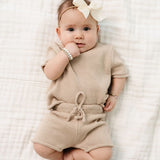 Baby with a white bow lying on a blanket in the Lou Lou & Company Knit Shorts and Tee in Tan