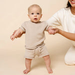 Baby standing with help wearing the Lou Lou & Company Knit Shorts and Tee in Tan