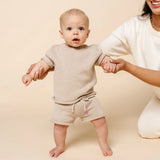Baby standing with help wearing the Lou Lou & Company Knit Shorts and Tee in Tan