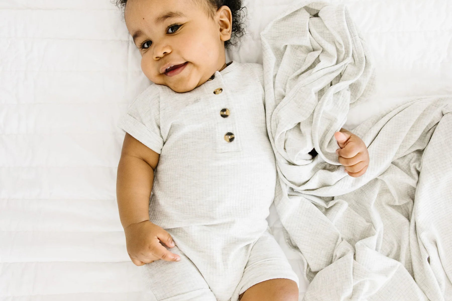 Baby lying on a white bed wearing the Lou Lou & Company Ribbed Romper in Stevie gray