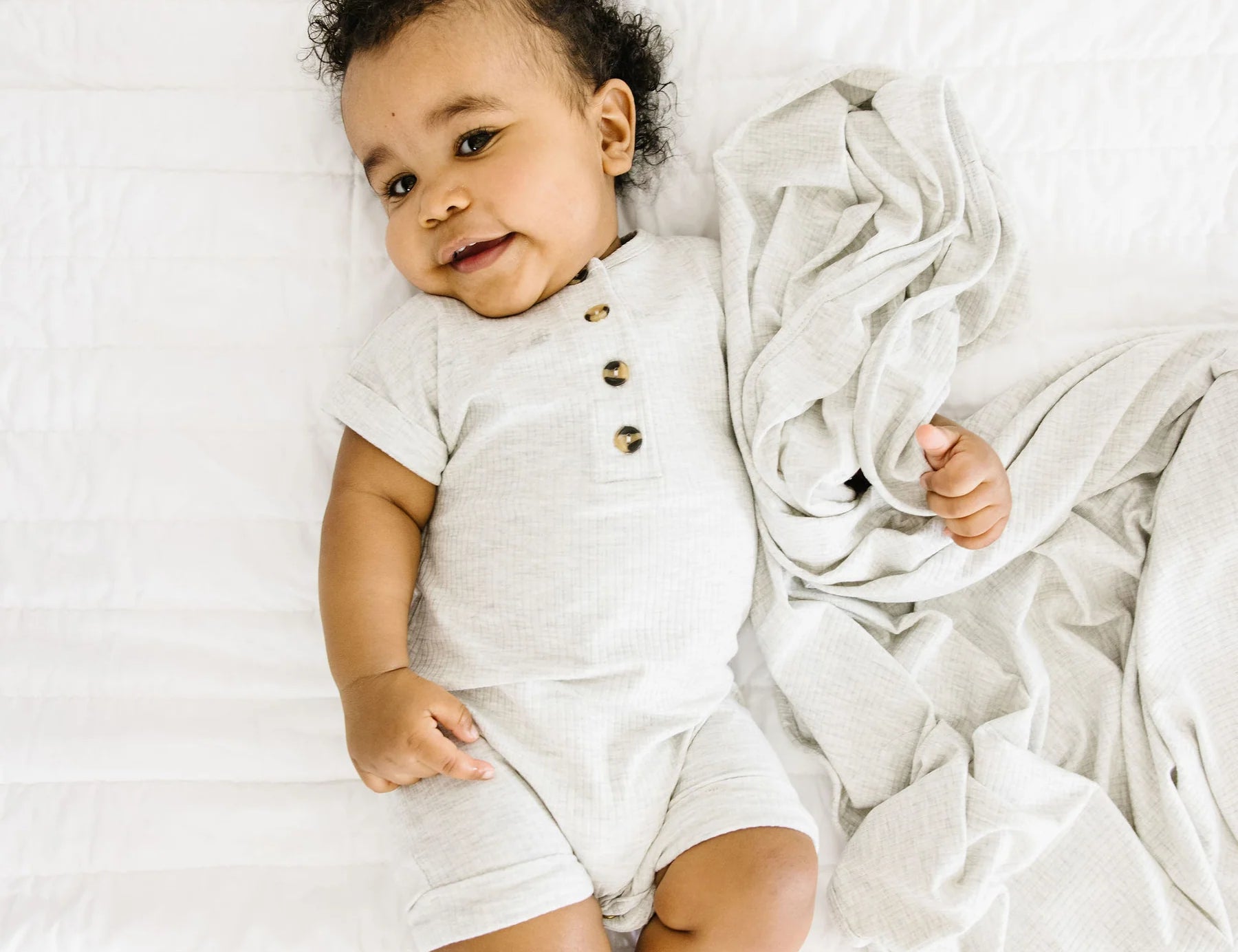Baby lying on a white bed wearing the Lou Lou & Company Ribbed Romper in Stevie gray