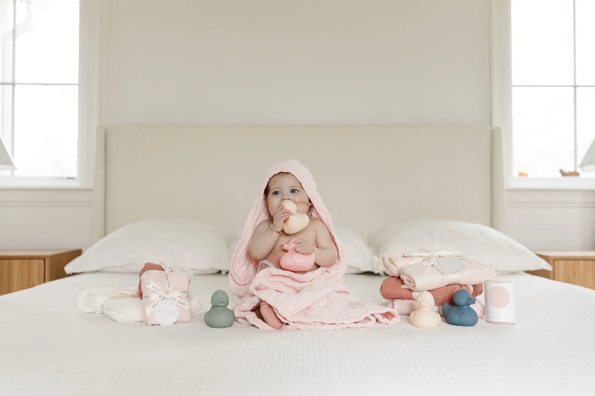 Baby in a pink hooded towel sitting on a bed with the Lou Lou & Company Rubber Duckies Set