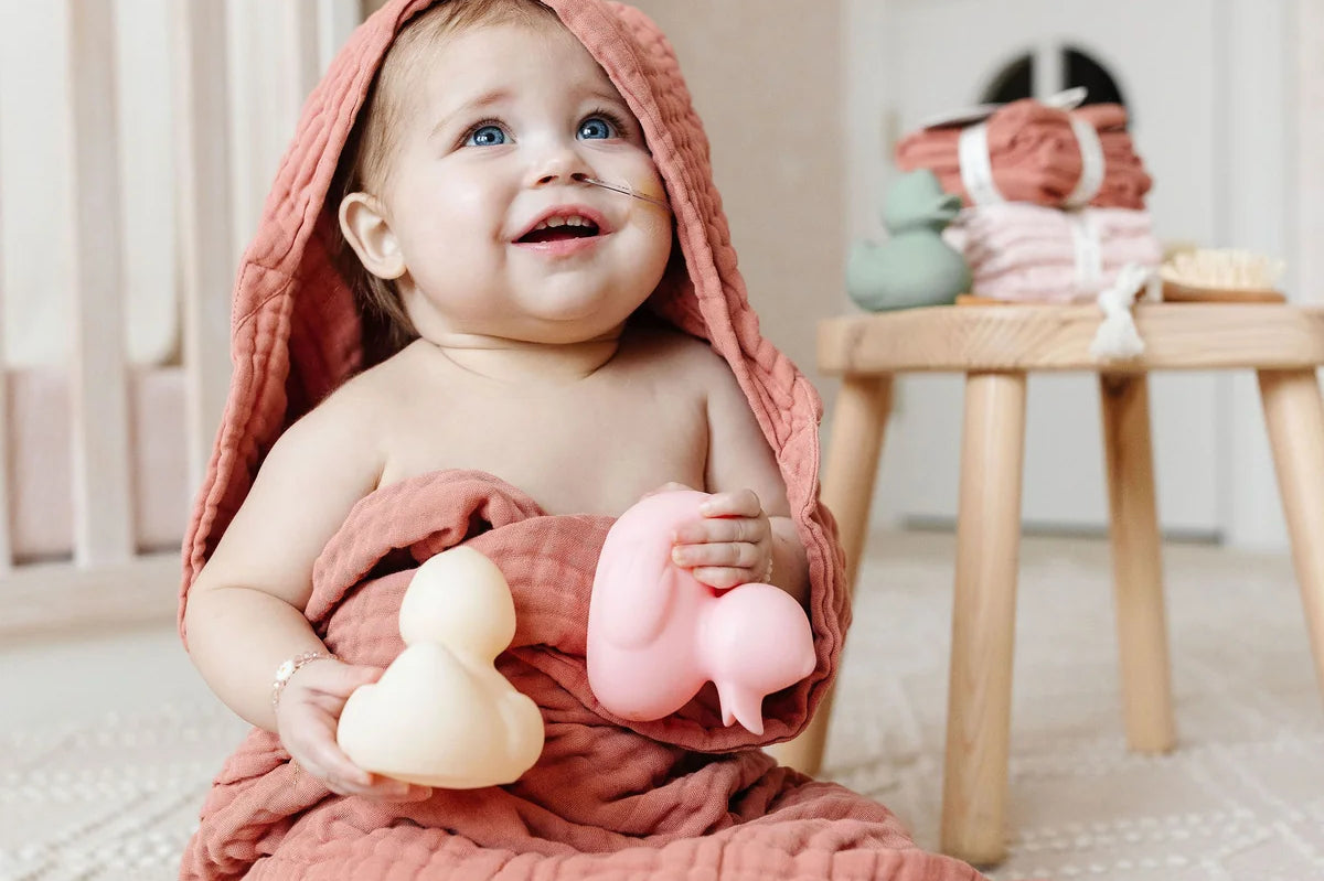 Smiling baby in a pink hooded towel holding Lou Lou & Company Rubber Duckies in a nursery