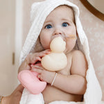 Baby in a hooded towel chewing on a cream Lou Lou & Company Rubber Ducky and holding a pink one