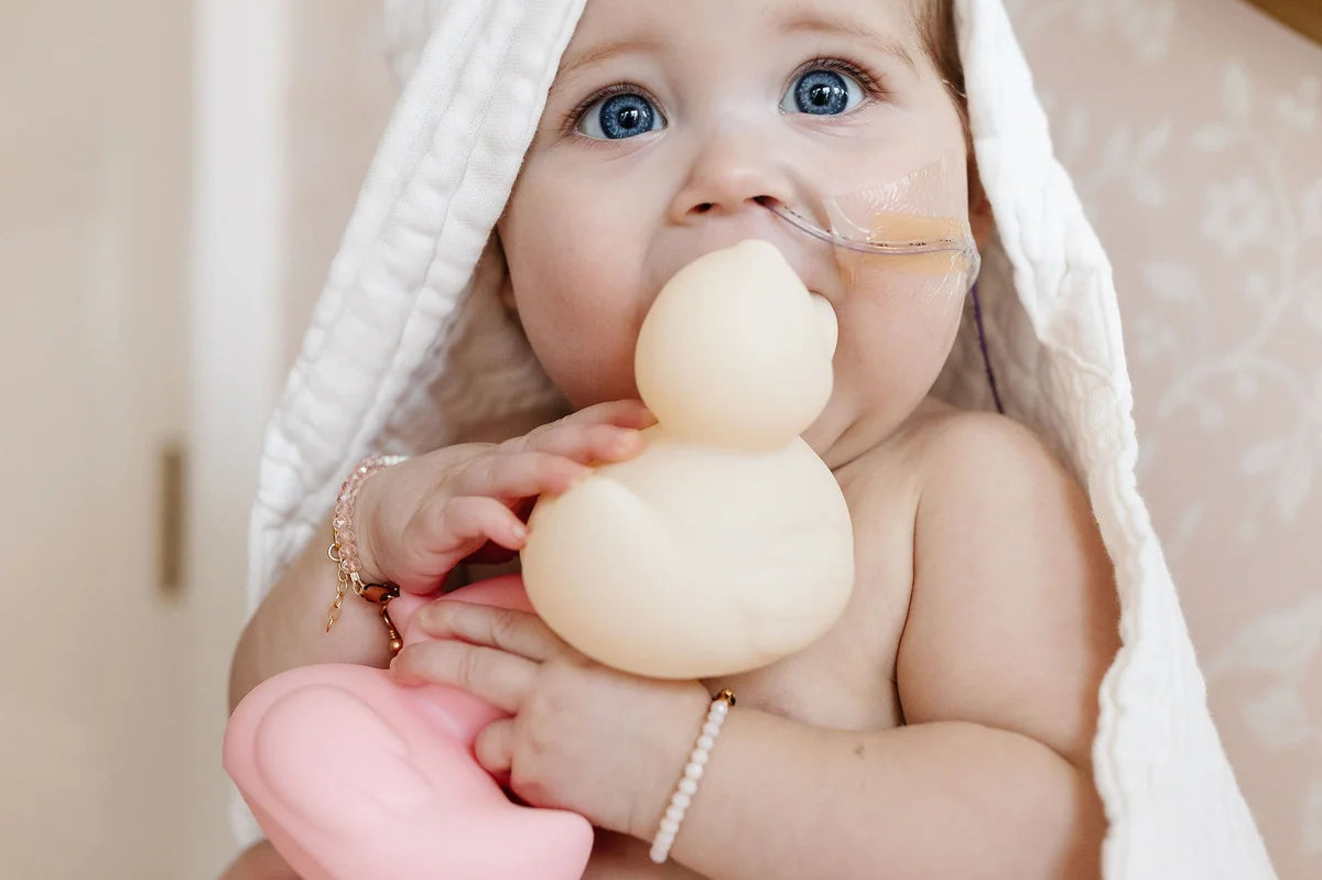 Baby in a hooded towel chewing on a cream Lou Lou & Company Rubber Ducky and holding a pink one