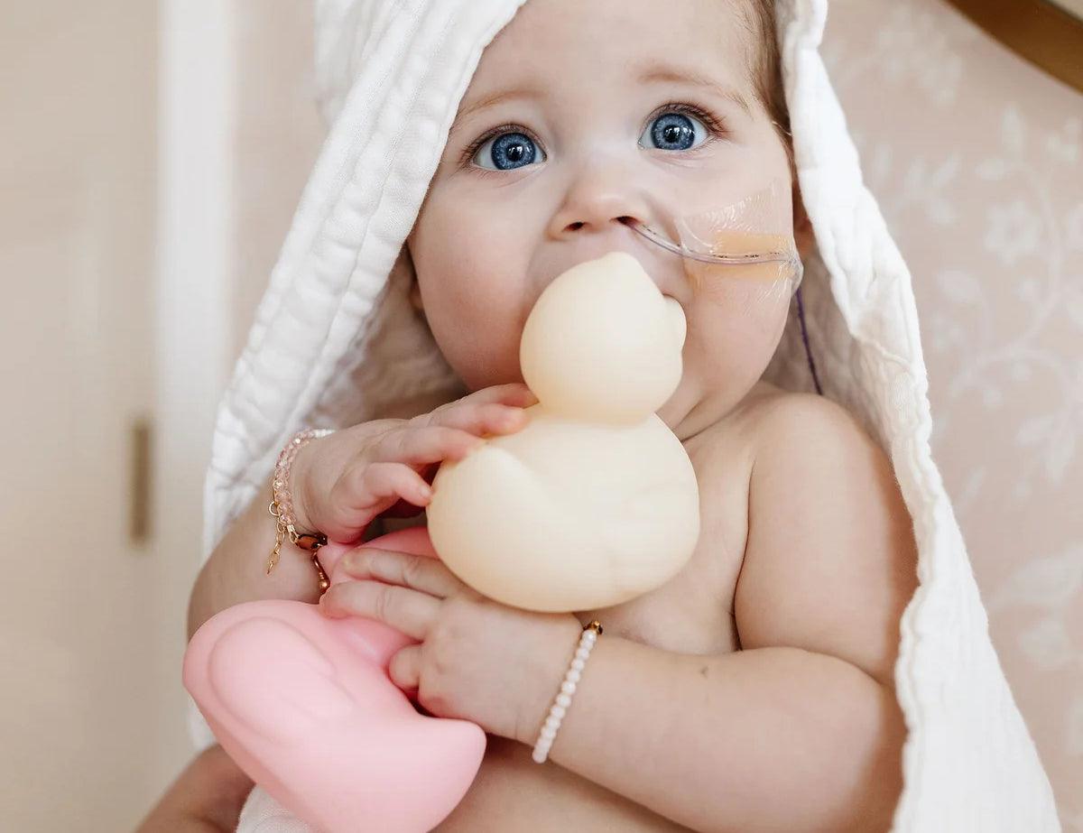 Baby in a hooded towel chewing on a cream Lou Lou & Company Rubber Ducky and holding a pink one