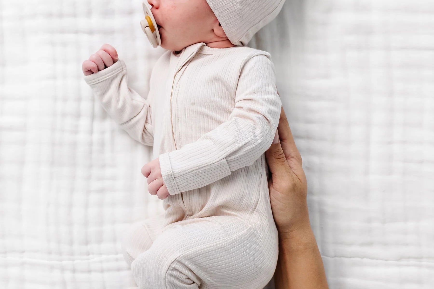 Newborn in a cream hat wearing the Lou Lou & Company Zipper Footies in Cove, lying on a muslin blanket