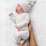 Newborn in a cream hat wearing the Lou Lou & Company Zipper Footies in Cove, lying on a muslin blanket