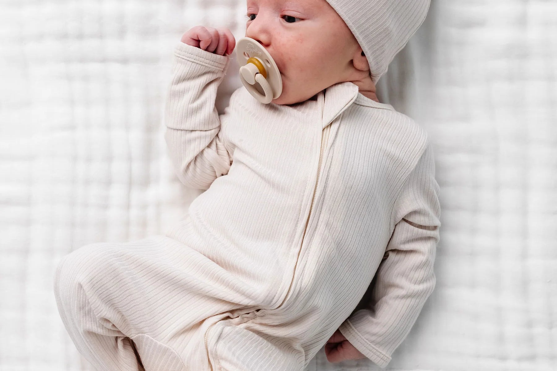 Newborn with a pacifier wearing the Lou Lou & Company Zipper Footies in Cove and a matching cream hat