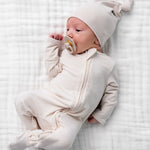 Newborn with a pacifier wearing the Lou Lou & Company Zipper Footies in Cove and a matching cream hat