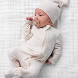 Newborn with a pacifier wearing the Lou Lou & Company Zipper Footies in Cove and a matching cream hat