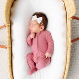 Newborn with a white bow sleeping in a bassinet wearing the Lou Lou & Company Zipper Footies in Marjoire, a mauve ribbed footie