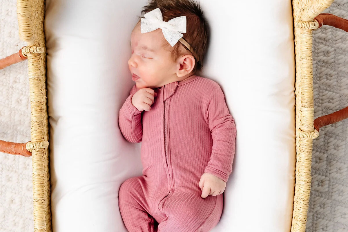 Newborn with a white bow sleeping in a bassinet wearing the Lou Lou & Company Zipper Footies in Marjoire, a mauve ribbed footie