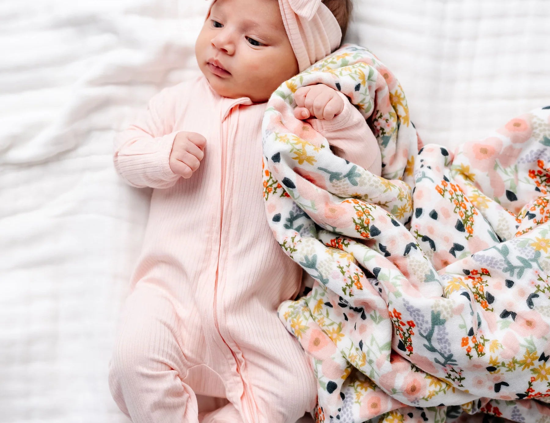 Newborn wearing the Lou Lou & Company Zipper Footies in Rosie, a light pink footie, beside a Bae floral blanket