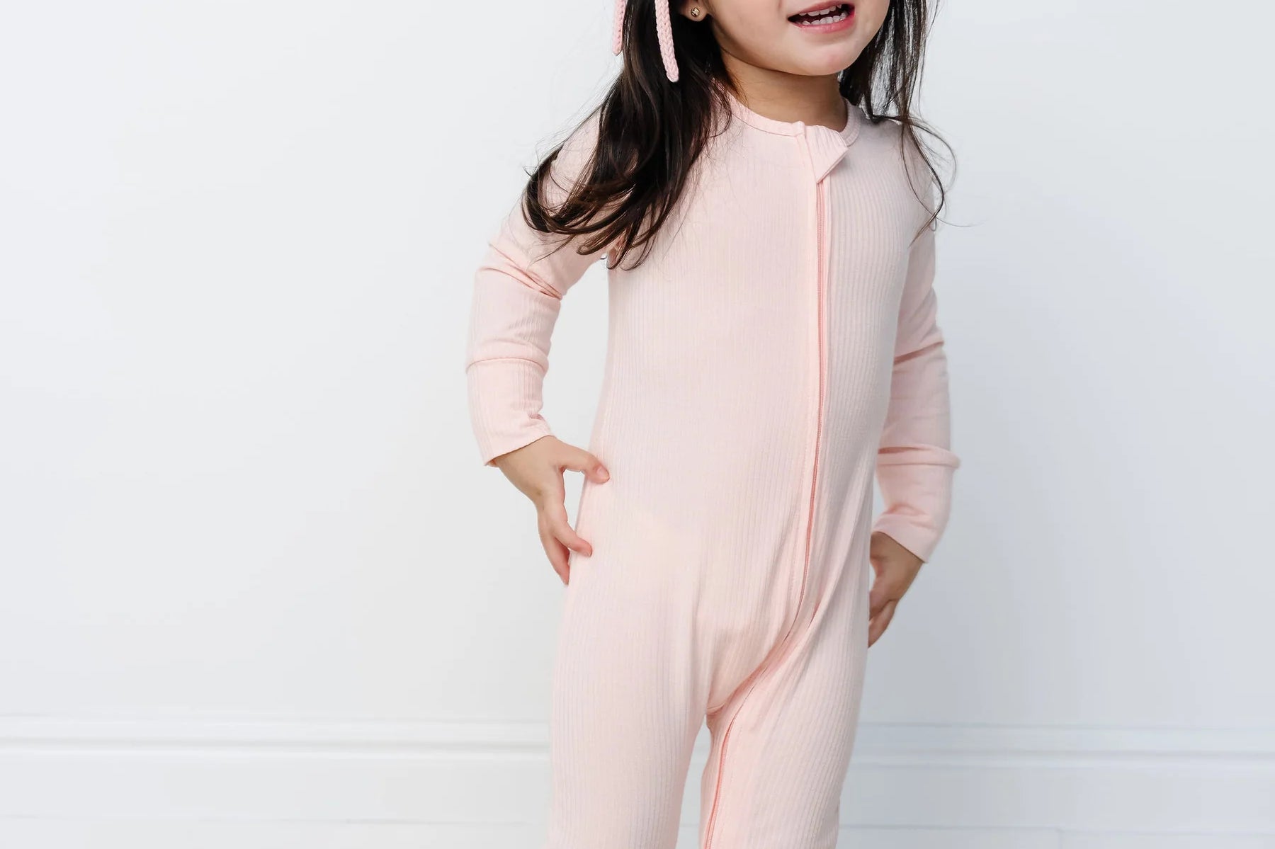 Toddler standing and wearing the Lou Lou & Company Zipper Footies in Rosie, a light pink ribbed footie