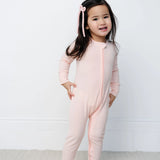 Toddler standing and wearing the Lou Lou & Company Zipper Footies in Rosie, a light pink ribbed footie