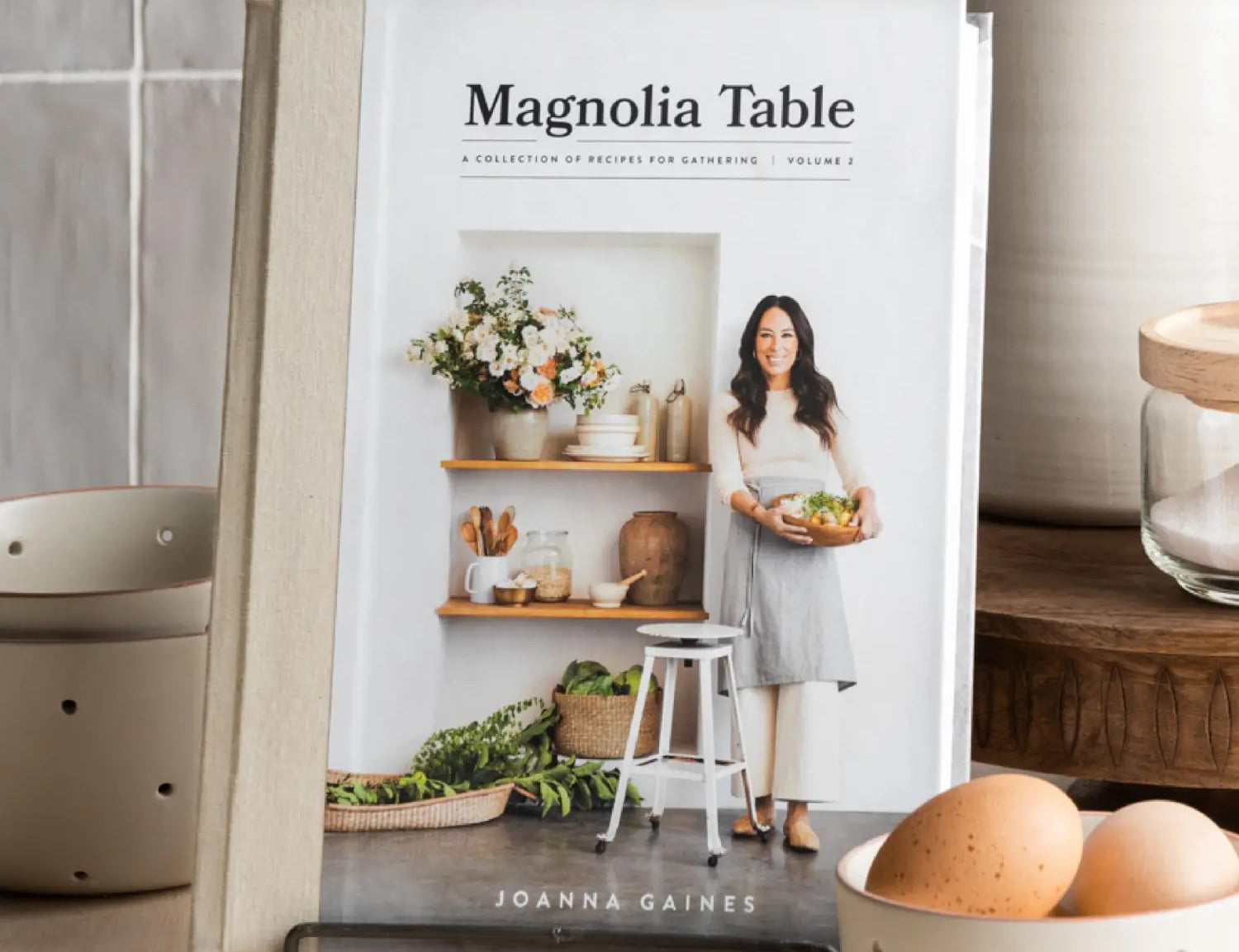 Magnolia Table Cookbook Volume 2 by Joanna Gaines on a stand in a kitchen with eggs and ceramics