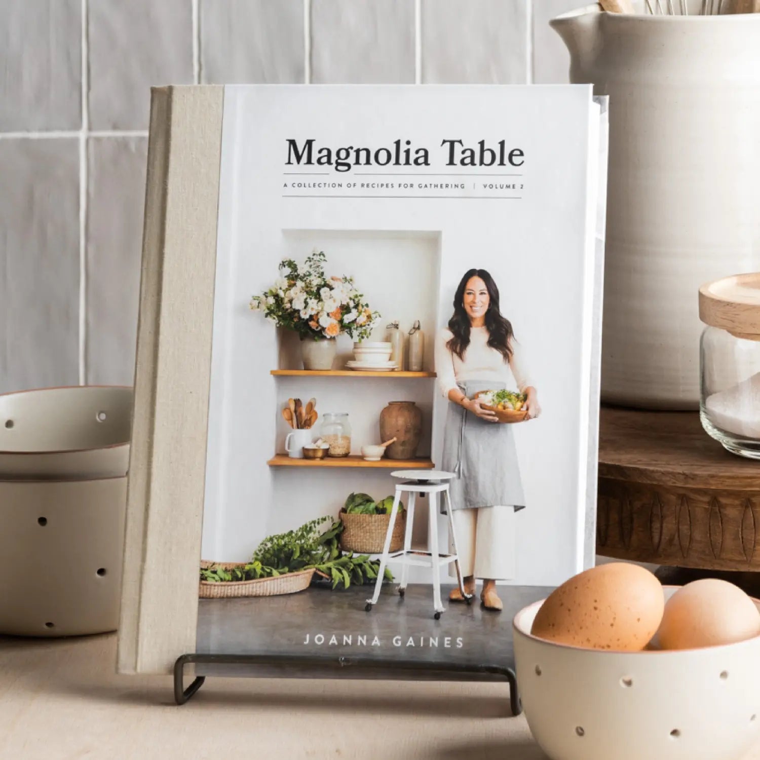 Magnolia Table Cookbook Volume 2 by Joanna Gaines on a stand in a kitchen with eggs and ceramics