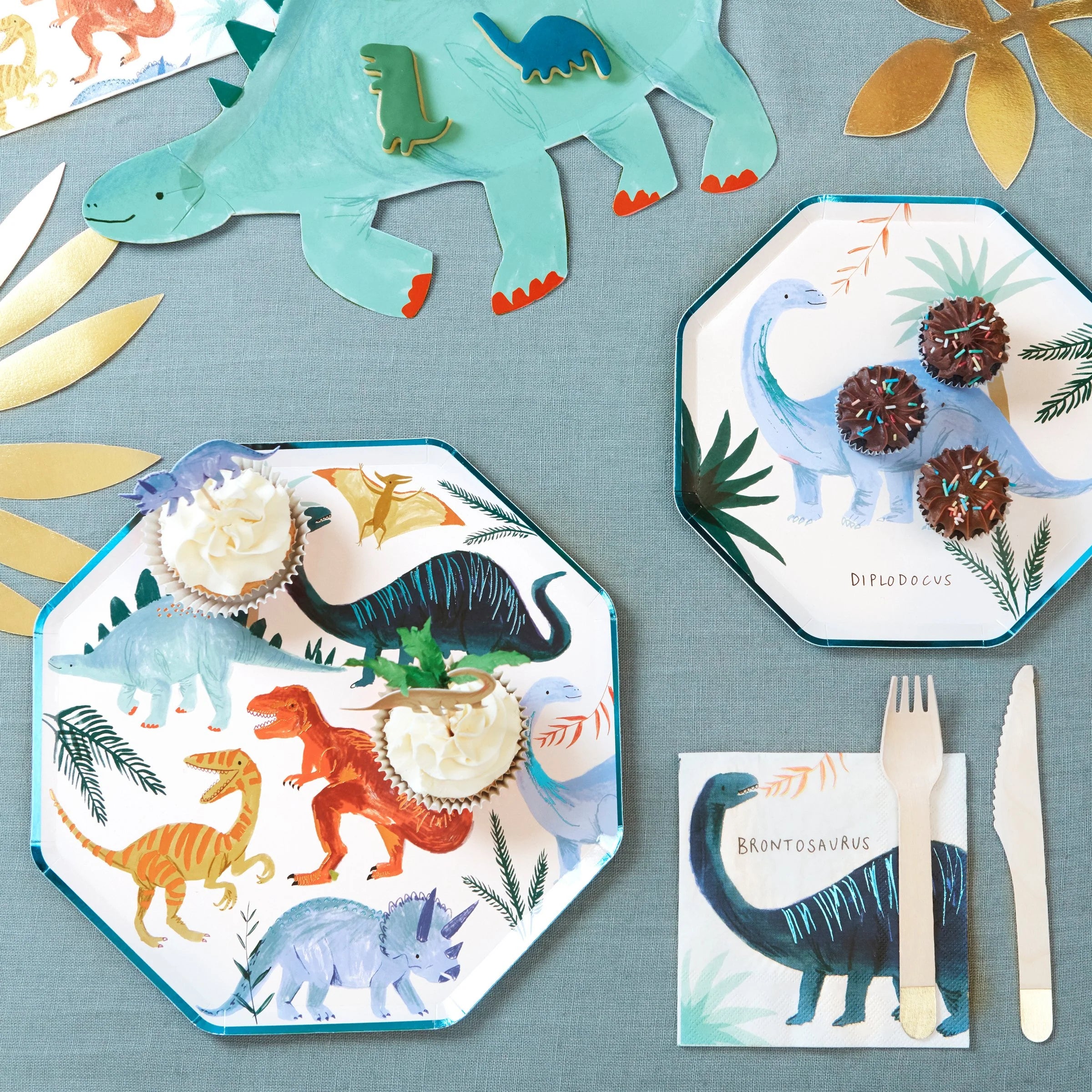 Meri Meri Dinosaur Kingdom Dinner Plates set on a teal tablecloth with cupcakes and dinosaur napkins
