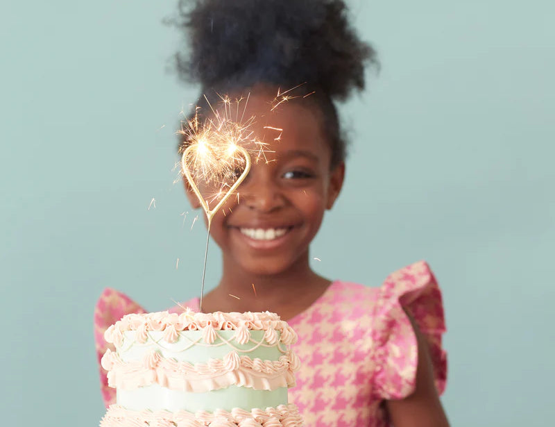 Smiling girl in a pink dress holding a cake lit with a Meri Meri Gold Sparkler Number Candle