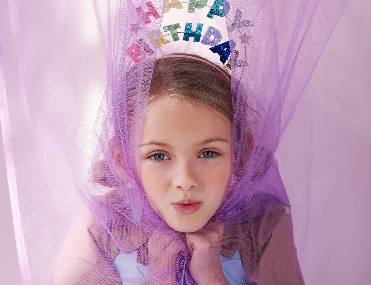Girl under a purple tulle canopy wearing the Meri Meri Happy Birthday Glitter Headband