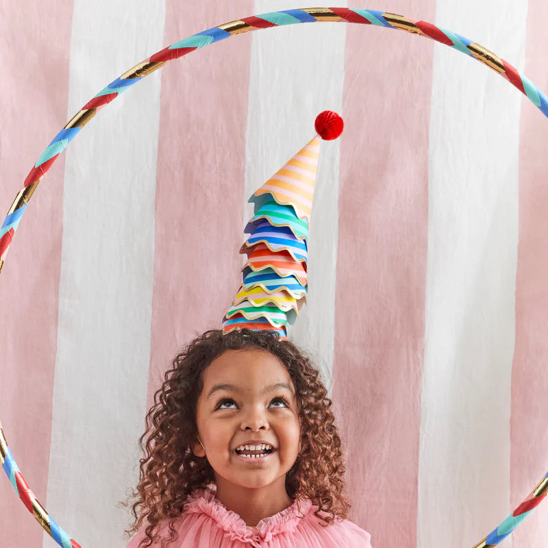 Smiling girl wearing a Meri Meri Stripe Party Hat under a rainbow hoop at a party