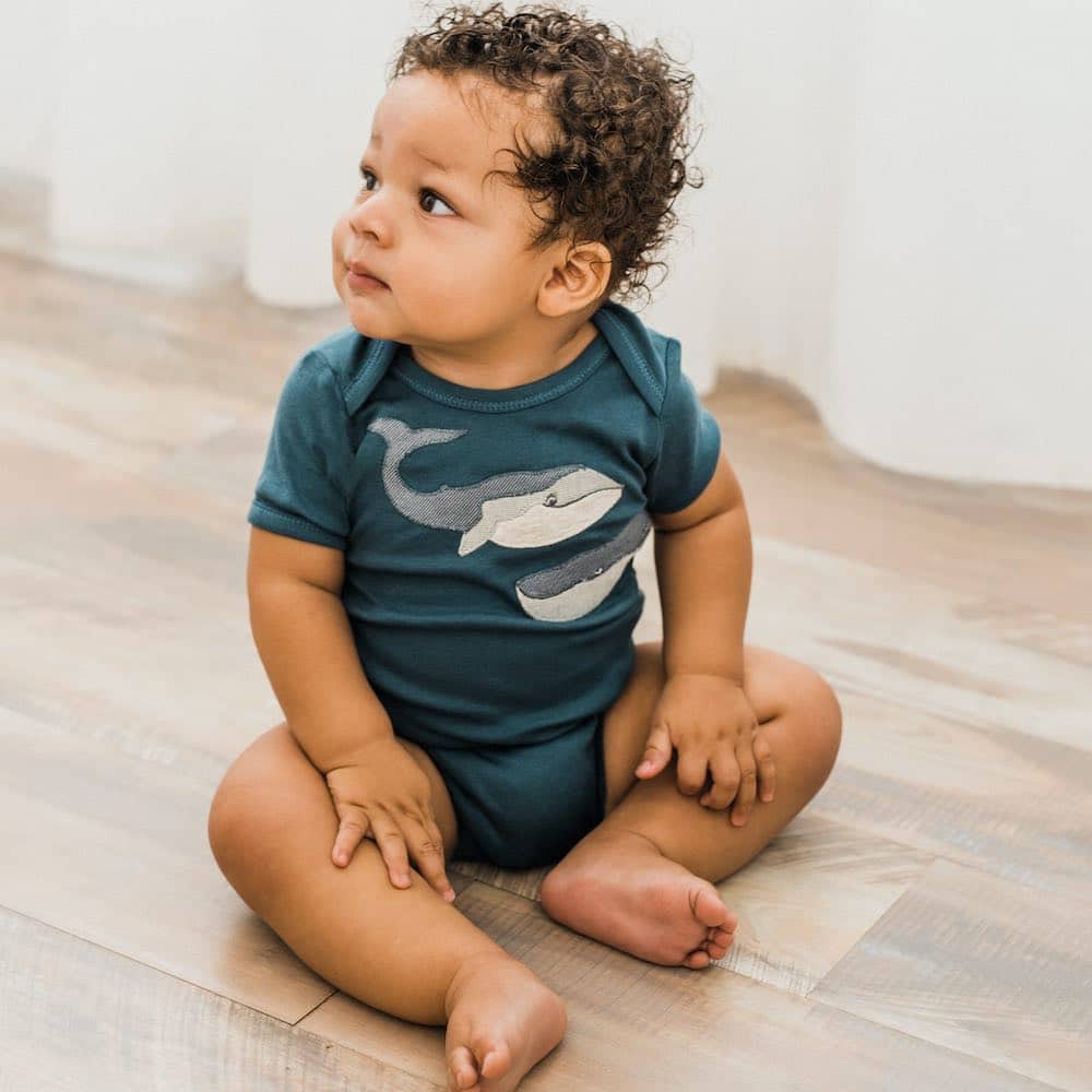 Baby with curly hair sitting on a wood floor wearing the Milkbarn Blue Whale Short Sleeve One Piece