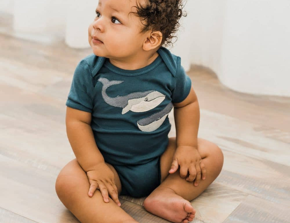 Baby with curly hair sitting on a wood floor wearing the Milkbarn Blue Whale Short Sleeve One Piece