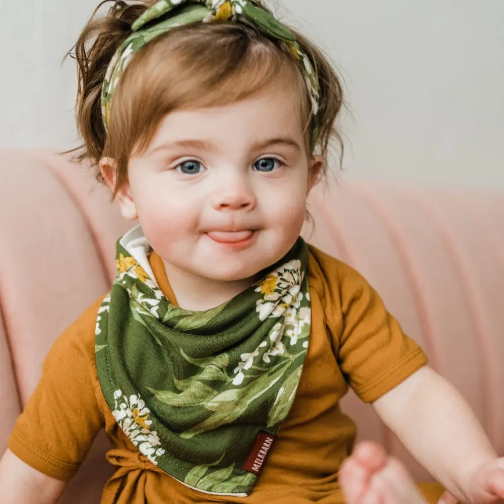 Smiling baby in a rust top wearing the Milkbarn Green Floral Three-Layer Kerchief Bib and matching headband