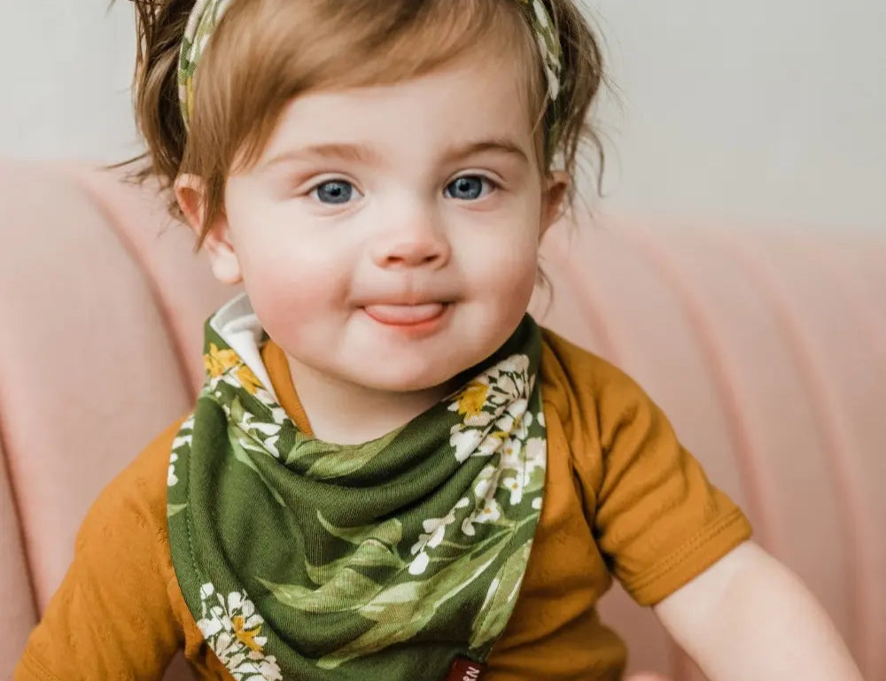 Smiling baby in a rust top wearing the Milkbarn Green Floral Three-Layer Kerchief Bib and matching headband