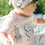 Baby in a floral headband wearing the Milkbarn Tutu Elephant Short Sleeve One Piece outdoors