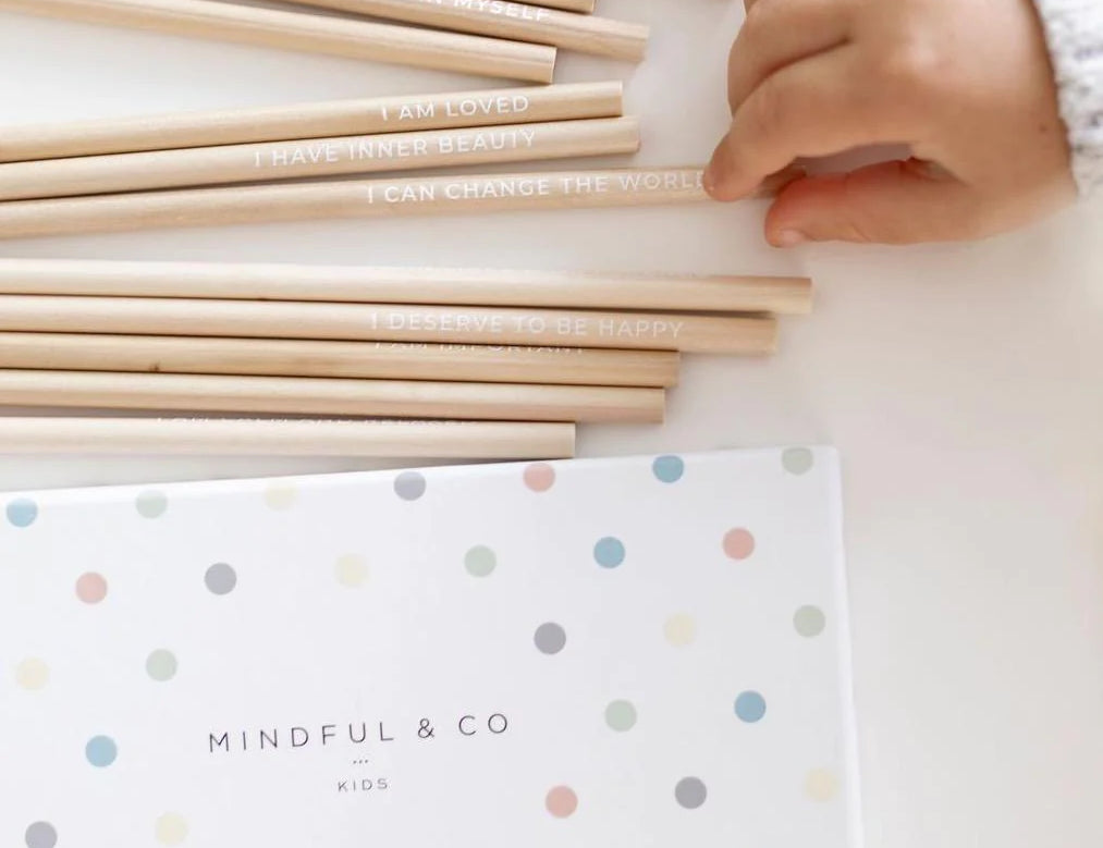 Mindful & Co Affirmation Coloring Pencils, a child's hand picking up a natural wood pencil beside the polka dot box