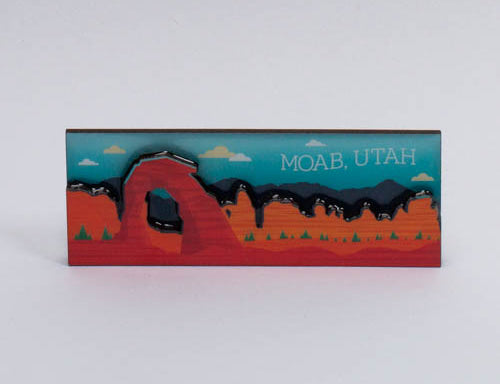 Layered Moab magnet with Delicate Arch, a dark ridge and pines cut in stacked depth under a teal sky