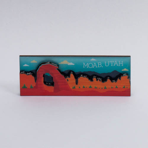 Layered Moab magnet with Delicate Arch, a dark ridge and pines cut in stacked depth under a teal sky
