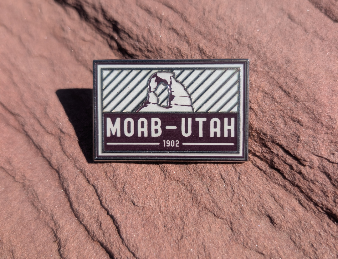 Gray and maroon Moab Utah 1902 enamel pin with Delicate Arch above diagonal stripes, lying on red sandstone in Moab, Utah.